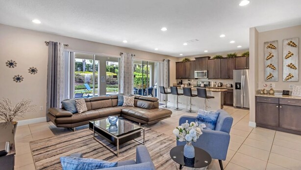 A modern living room with a sectional sofa, a coffee table, and a blue armchair. The kitchen is visible with dark cabinets and stainless steel appliances. - 7462 Modern 6BR/6BA Pool Home near Disney (Kissimmee)