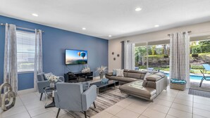 A modern living room with a sectional sofa, a flat-screen TV, and a dining area. - 7462 Modern 6BR/6BA Pool Home near Disney (Kissimmee)