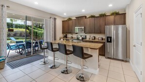 A modern kitchen with a central island, bar stools, a refrigerator, and a view of a pool and garden. - 7462 Modern 6BR/6BA Pool Home near Disney (Kissimmee)