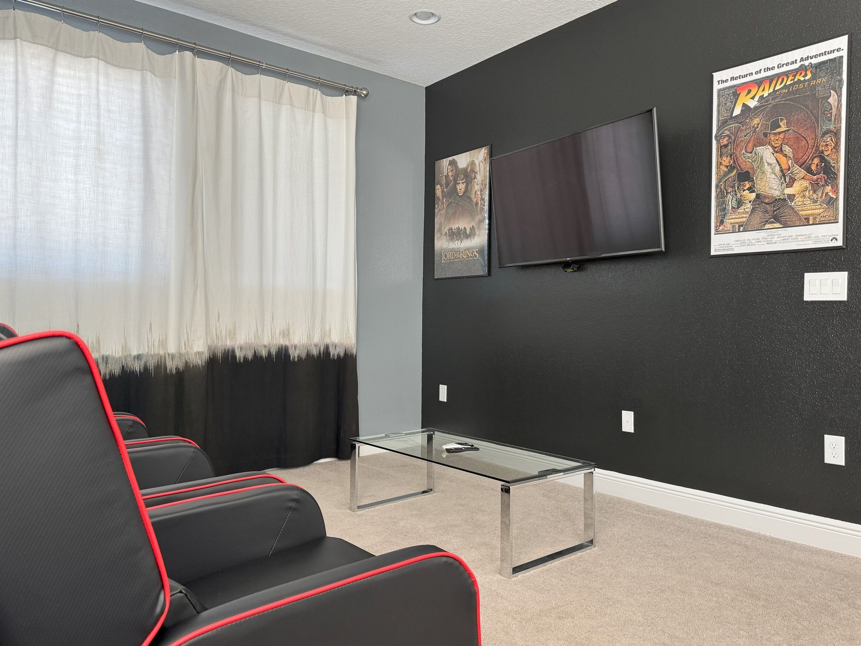 A room with black walls, a flat-screen TV, two posters, a glass coffee table, and black and red upholstered chairs.