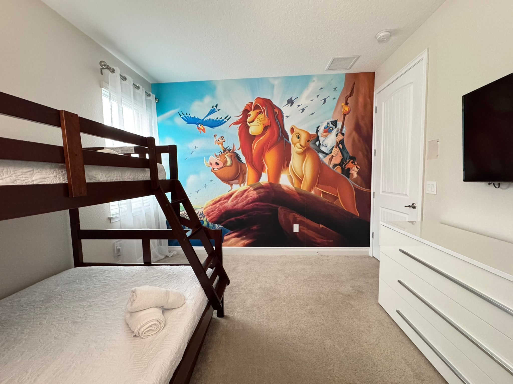 Un dormitori amb un mural de personatges animats, una llitera, un televisor i un prestatge blanc.