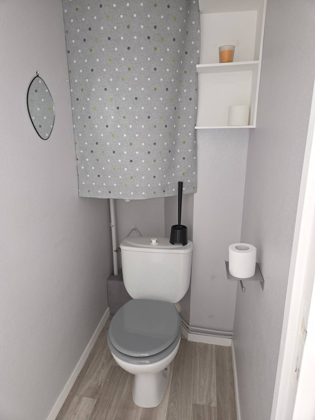 A small bathroom with a toilet, a toilet paper holder, and a shelf with a cup and a roll of toilet paper.