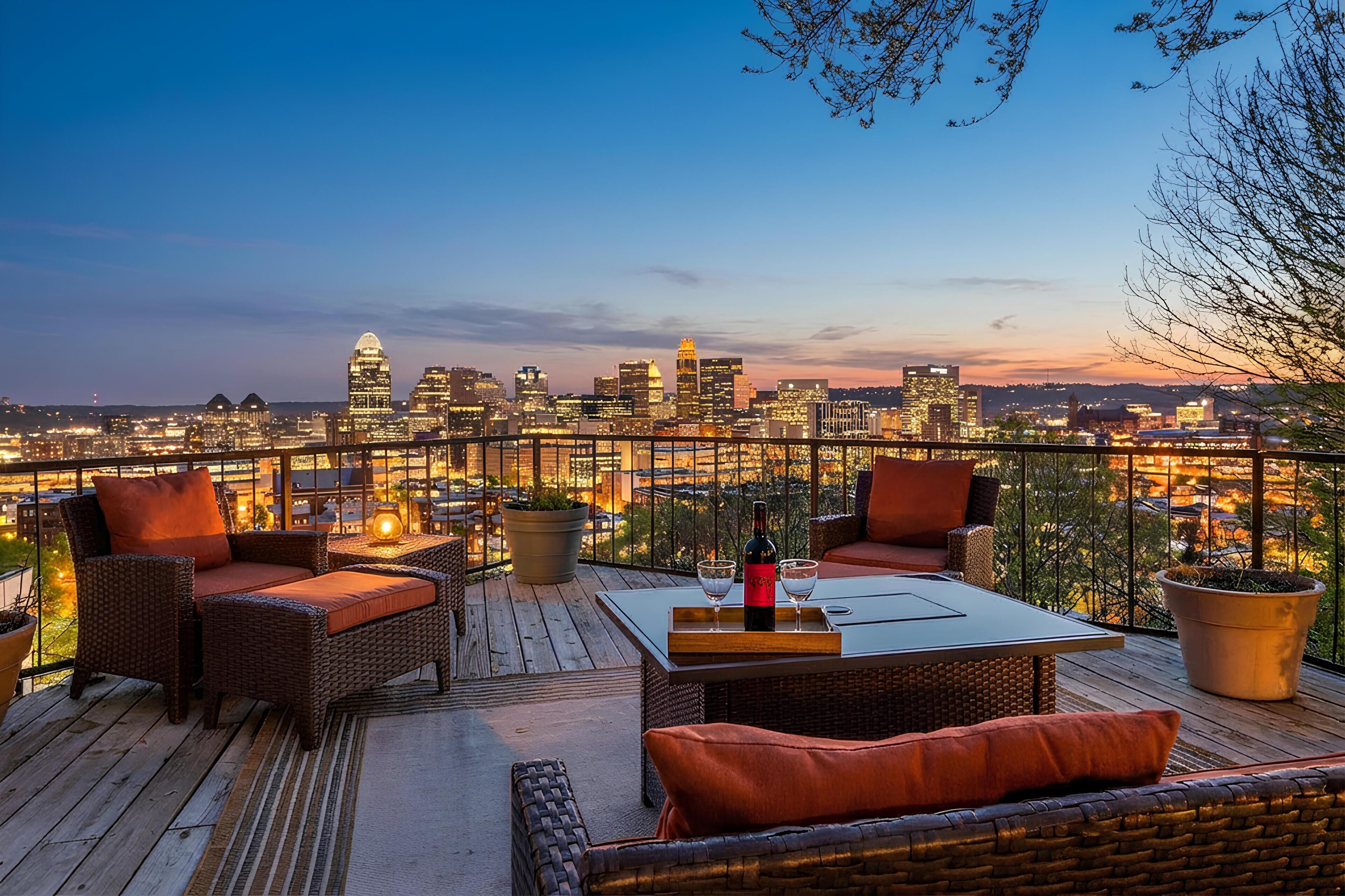 A rooftop terrace with wicker furniture, a table with a bottle and glasses, and a city skyline view.