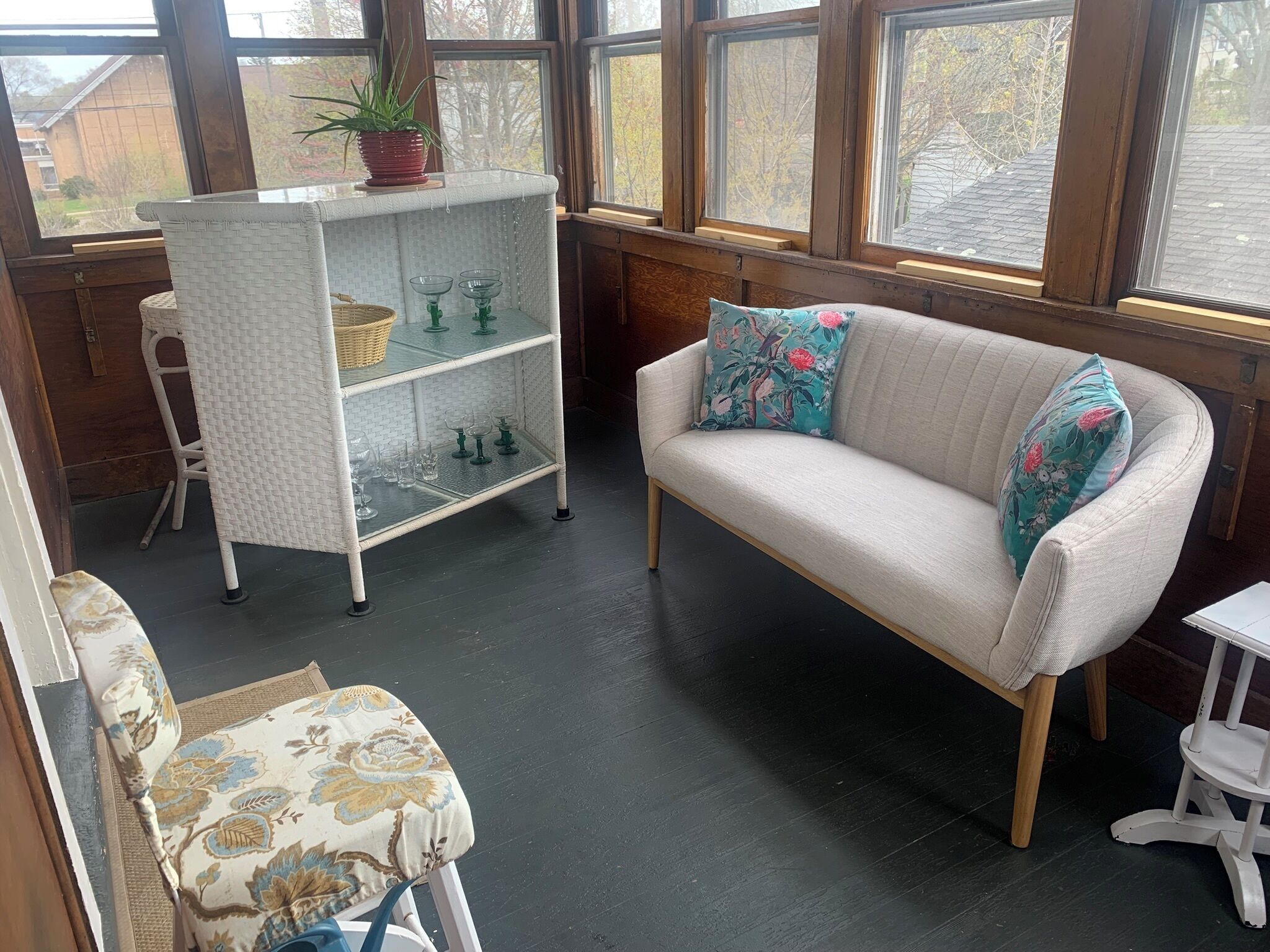 Een kamer met een witte bank, een rieten bijzettafeltje met een plant, een glazen kast met planken en uitzicht door grote ramen.
