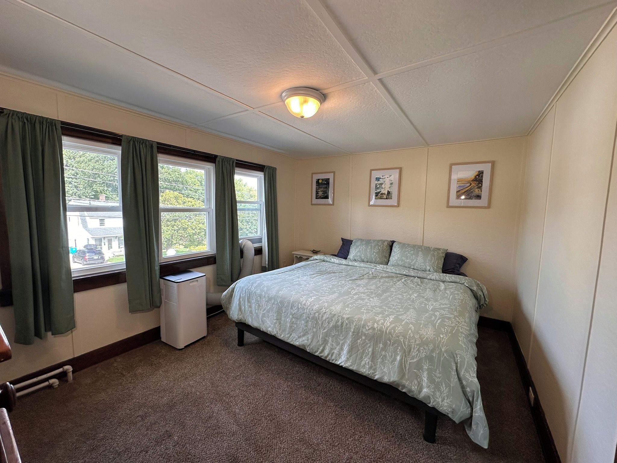 A bedroom with a bed, a window with curtains, a radiator, and framed pictures on the wall.