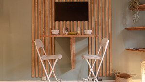 A room with a wooden slat wall, a flat-screen TV, two white chairs, a small table with a bowl and a candle, and a wicker basket. - Studio Victoria - Welkeys (Biarritz)