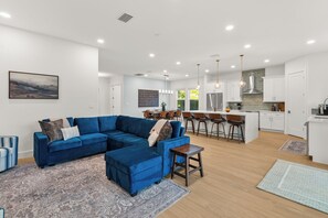 Ein modernes Wohnzimmer mit einem blauen Ecksofa, einem Essbereich mit Barhocker und einer Küche mit weißen Schränken und Edelstahlgeräten.