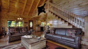 Ein geräumiges Blockhaus-Interieur mit zwei Ledersofas, einem Holztisch und einer Treppe, die zu einer oberen Ebene führt.