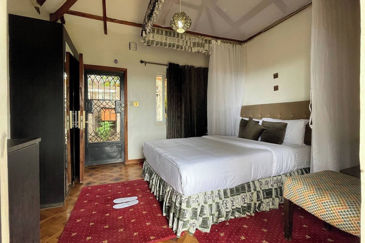Une chambre à coucher comprenant un lit, une table de chevet, une armoire, un tapis rouge à motifs, un ventilateur de plafond et une fenêtre avec des rideaux.