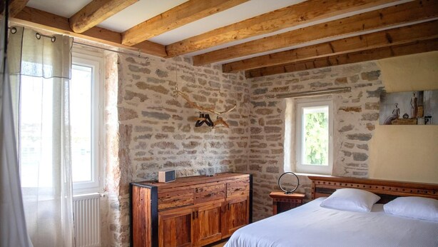 Camera da letto con parete in pietra, travi in legno, un letto, una cassettiera e una finestra vista sul verde.
