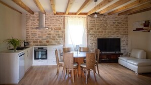 Un accogliente soggiorno con parete in pietra, travi in legno, zona pranzo con sedie, un divano e una TV.