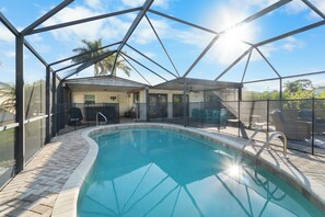 Piscina com estrutura de vidro, área de estar com cadeiras e cozinha externa coberta.