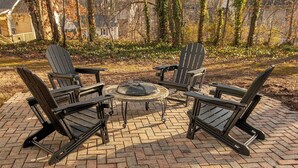 A brick patio with Adirondack chairs and a round table, surrounded by trees and houses. - McConnell Golf Retreat Near Sedgefield Country Club (Greensboro)