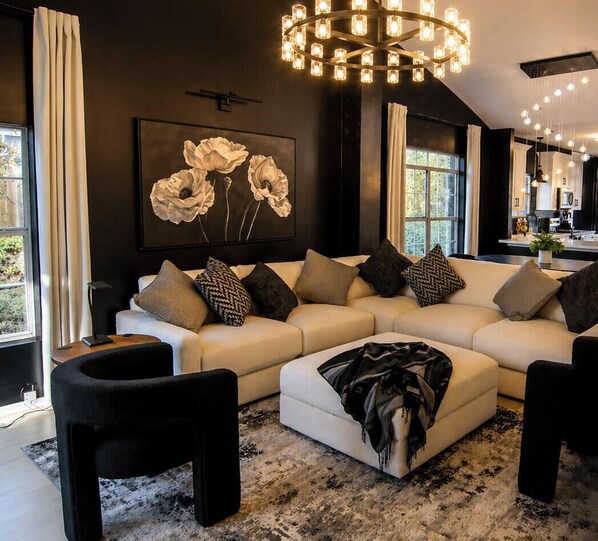 Un salon moderne avec un canapé d’angle, un pouf noir et un lustre.