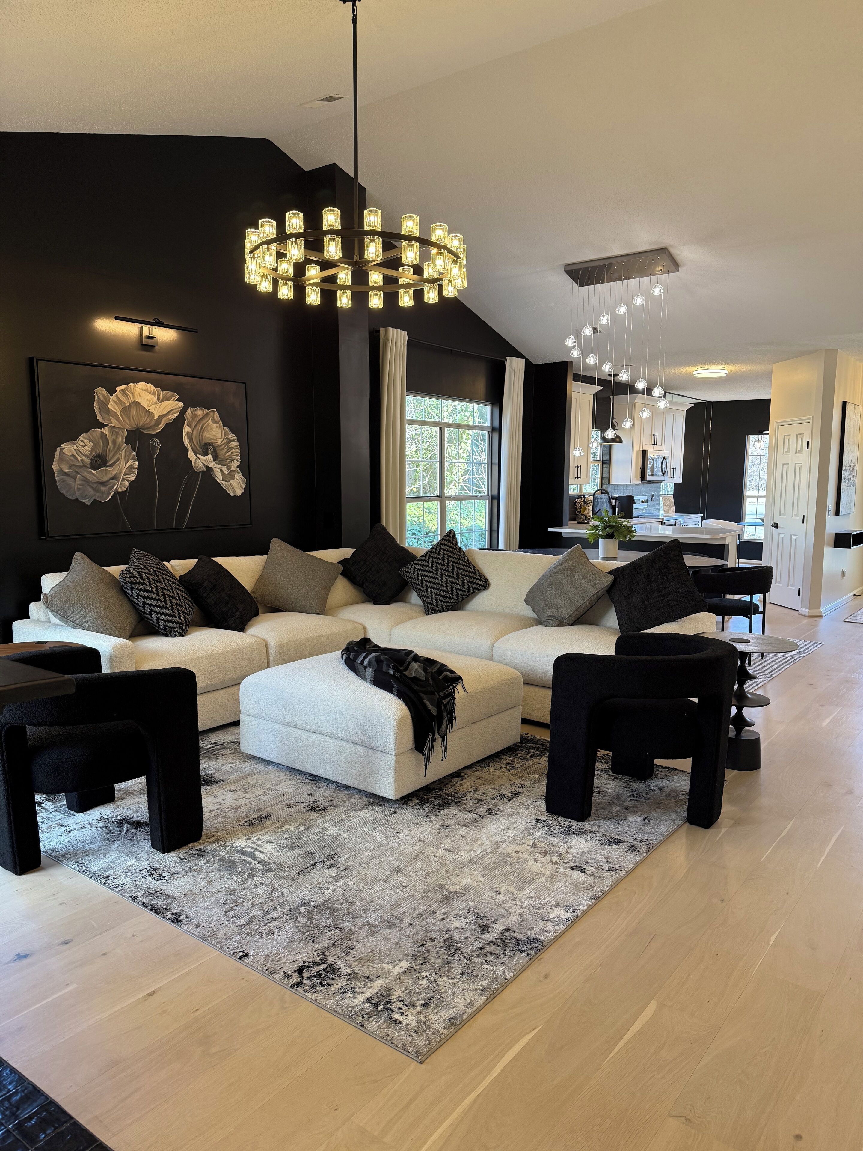 Ein modernes Wohnzimmer mit einem Ecksofa, einem schwarzen Couchtisch und einem großen Teppich. Im Hintergrund ist ein Essbereich mit einer sichtbaren Küche zu erkennen.