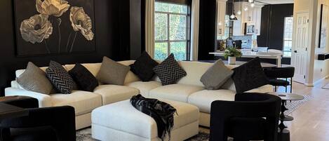 Ein modernes Wohnzimmer mit einem Ecksofa, einem schwarzen Couchtisch und einem großen Teppich. Im Hintergrund ist ein Essbereich mit einer sichtbaren Küche zu erkennen.