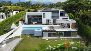 A modern two-story house with a swimming pool, surrounded by lush greenery and palm trees.