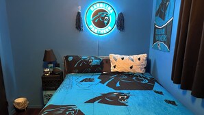 A bedroom with a Carolina Panthers themed bedspread, a neon sign, and a wall pillow. - Fully Furnished 3BR Home • Fast Wi-Fi • Parking • Near Uptown (Charlotte)