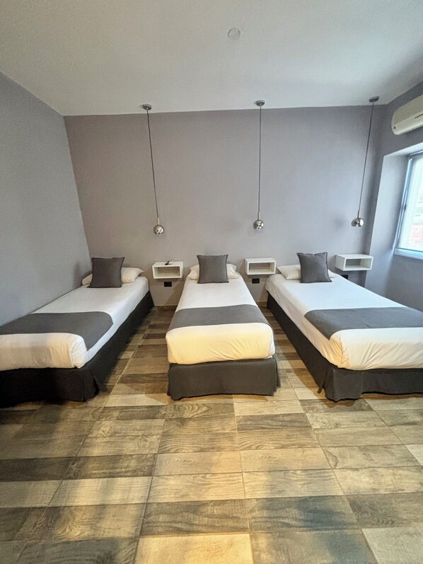 Habitación de hotel con tres camas individuales, una mesita de noche central y un ventanal con unidad de aire acondicionado.