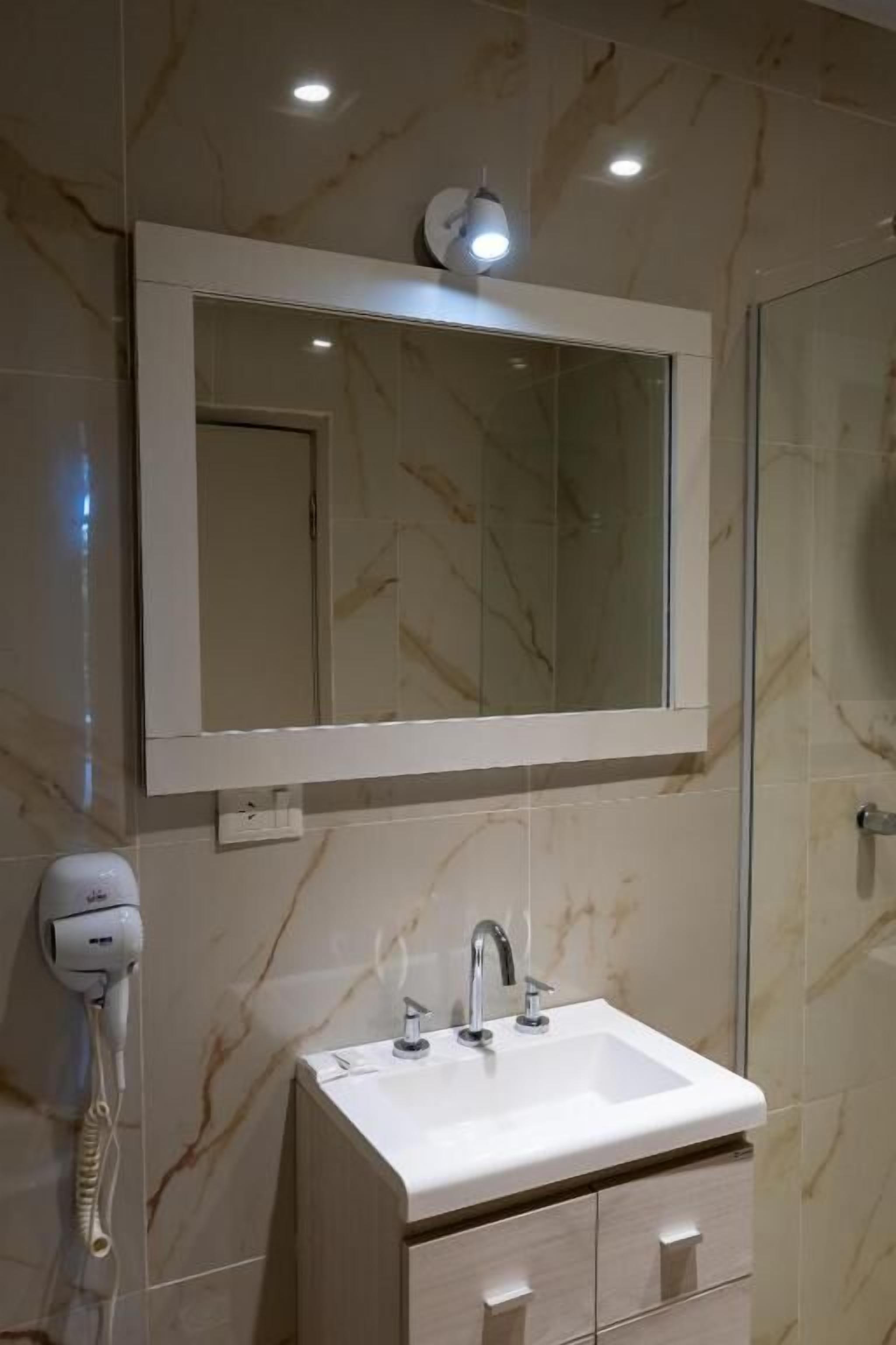 Une salle de bain avec un mur en marbre, un lavabo blanc avec un miroir au-dessus, et un sèche-cheveux fixé au mur.
