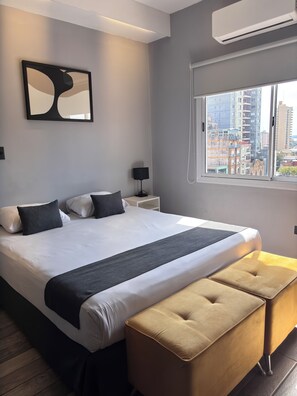 A modern hotel room with a large bed, a bedside table with a lamp, a window with a city view, and an air conditioning unit. - Hotel city (Posadas)