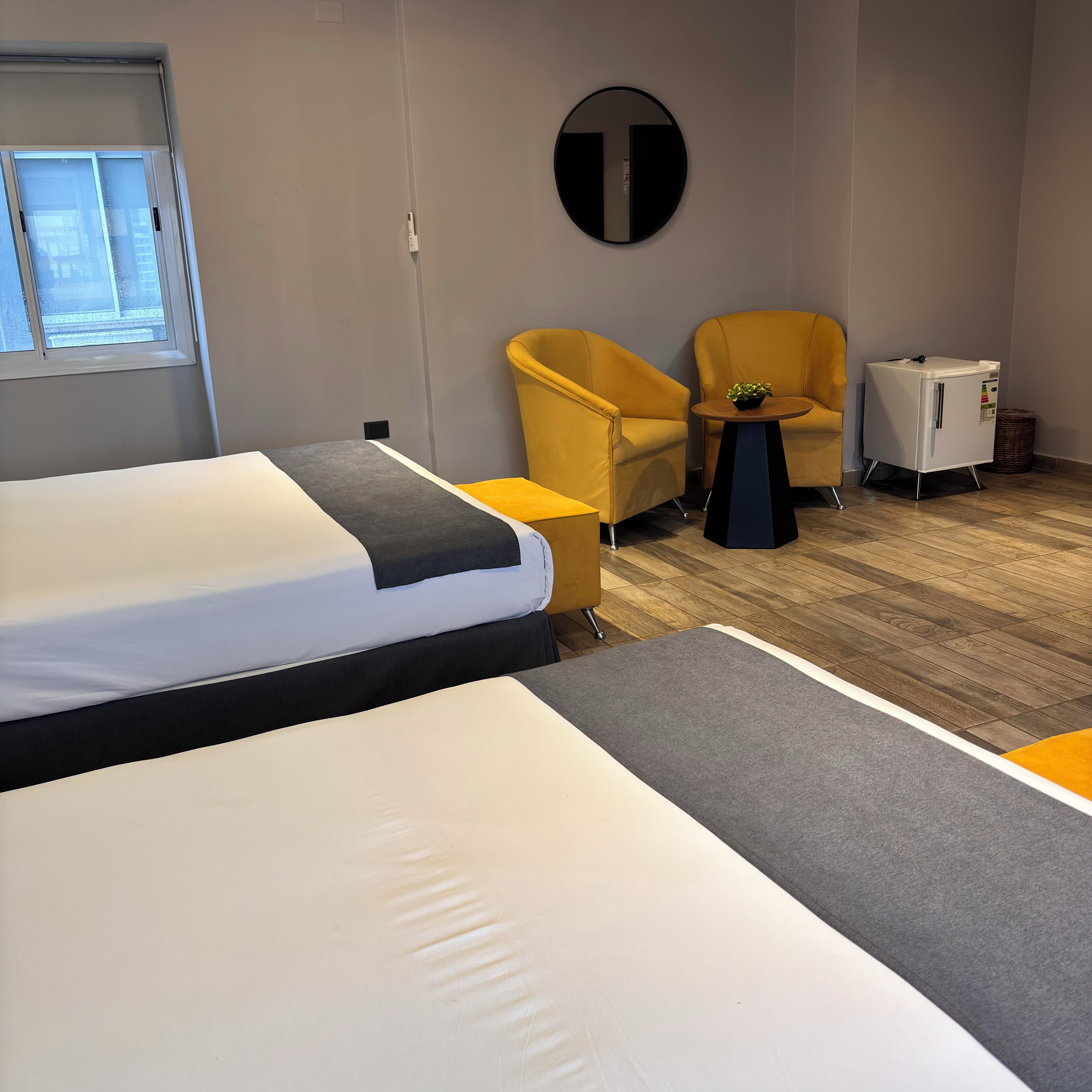 A hotel room with a bed, a yellow armchair, a small table with a plant, and a refrigerator.