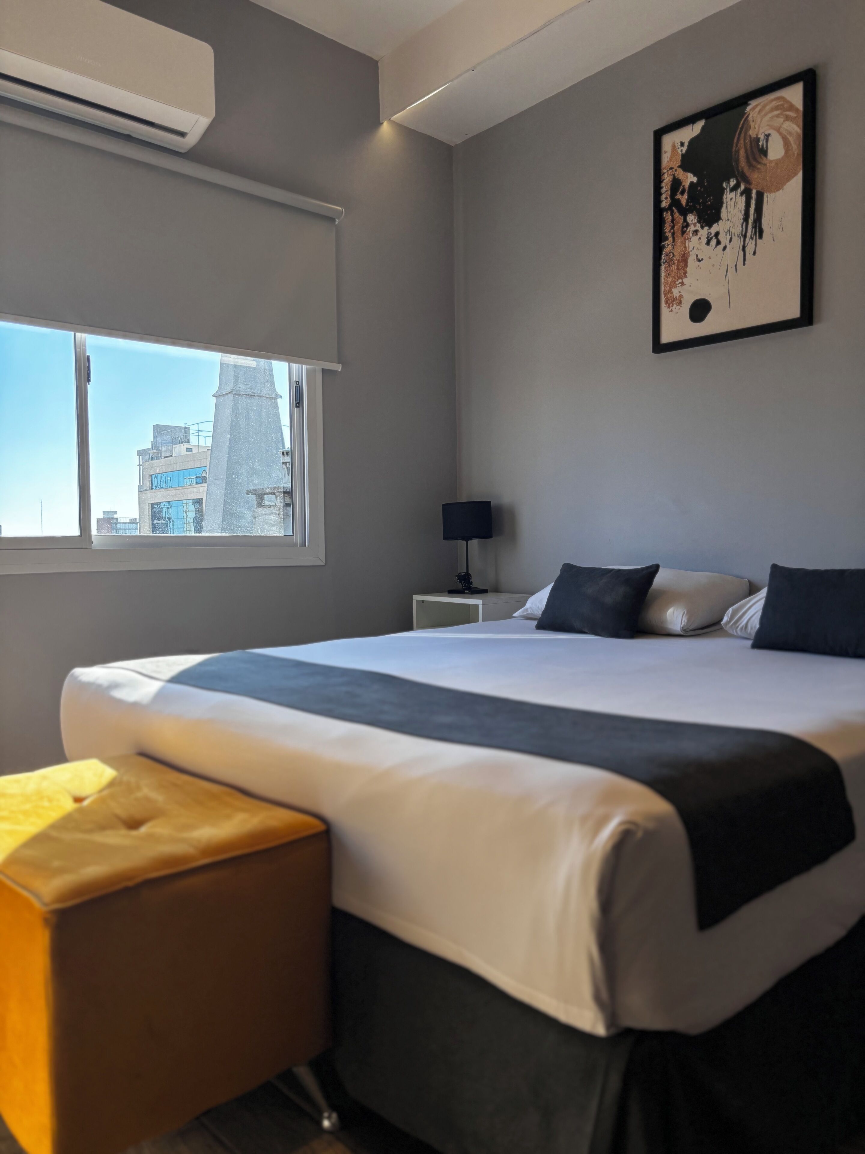 A modern hotel room with a large bed, a bedside table, a lamp, a window with a city view, and an air conditioning unit.