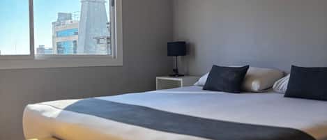 A modern hotel room with a large bed, a bedside table, a lamp, a window with a city view, and an air conditioning unit.