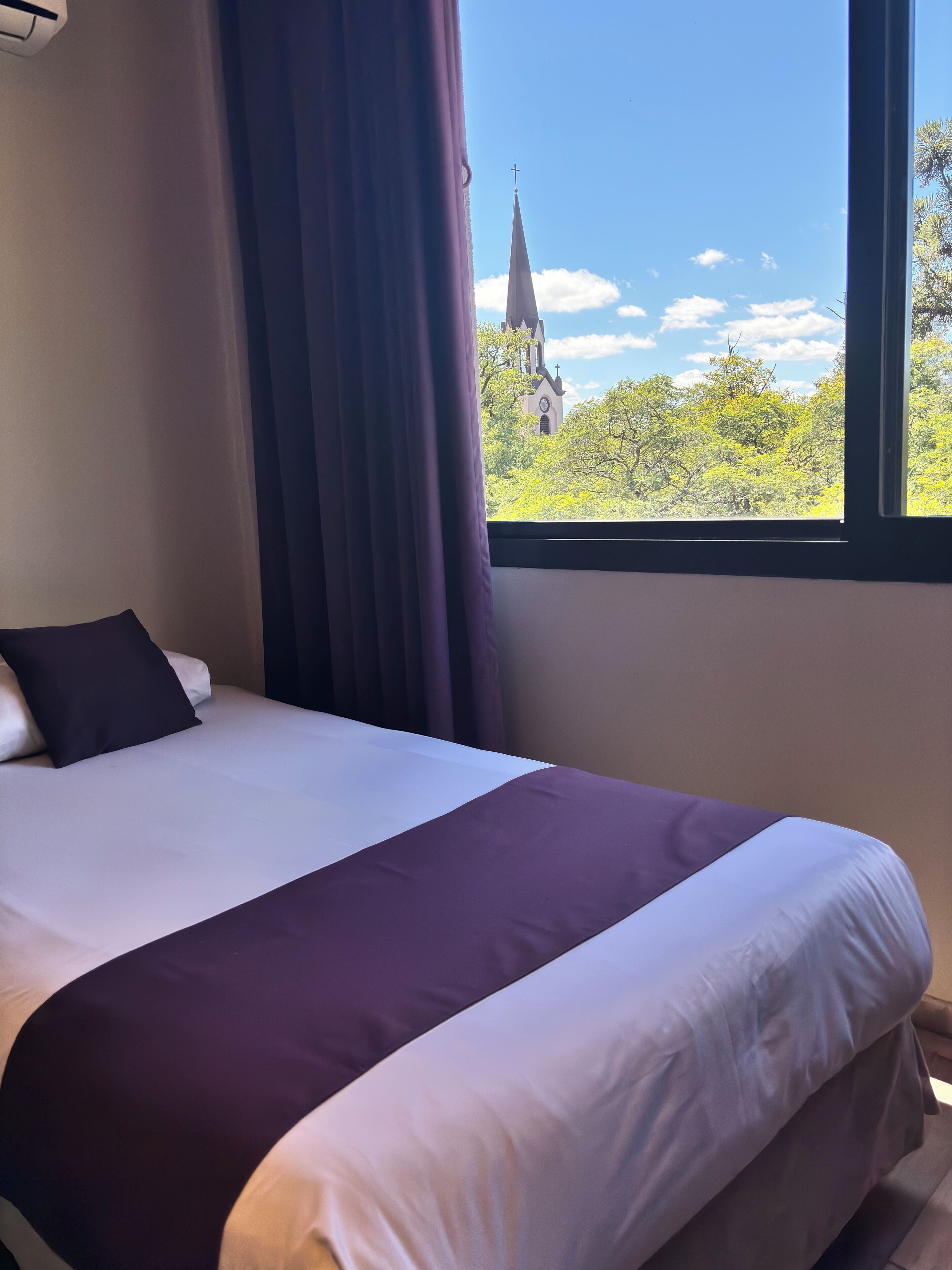 A neatly made bed with a white and purple bedspread, a dark pillow, and a window with a view of a church steeple and trees.