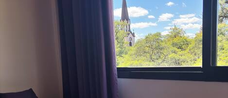 A neatly made bed with a white and purple bedspread, a dark pillow, and a window with a view of a church steeple and trees.