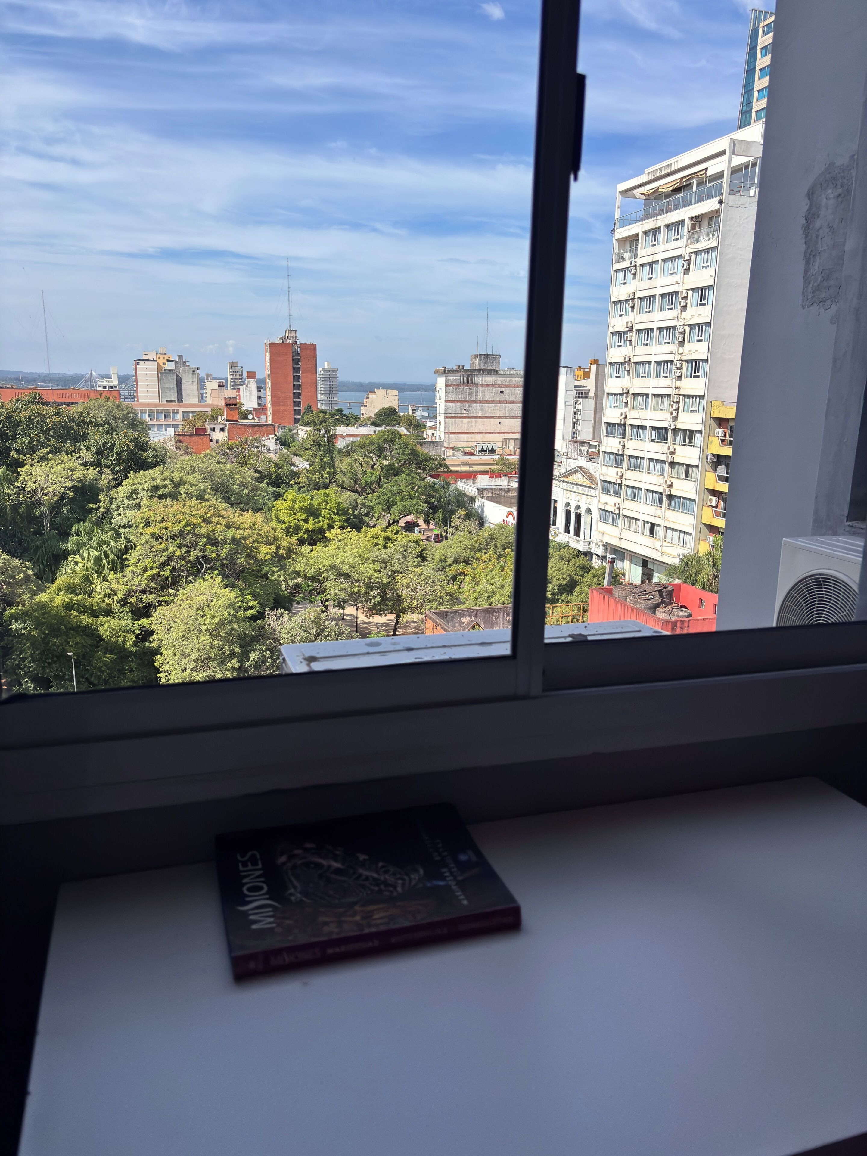 A room with a view of a cityscape through a window, a book on a table, and a white wall.