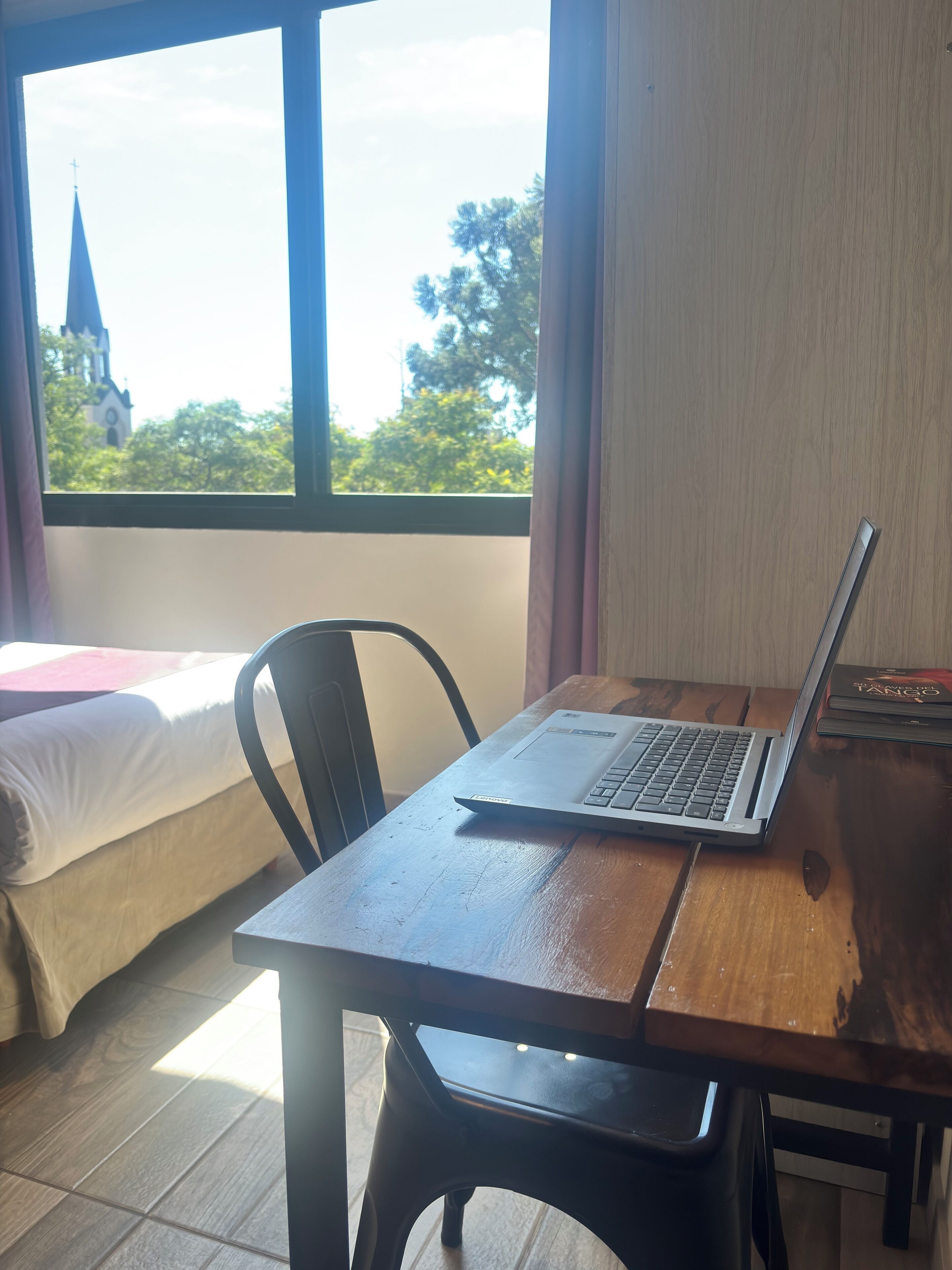 Una habitación con cama, un escritorio de madera con una computadora portátil, una silla y una ventana con vistas a una iglesia y árboles.