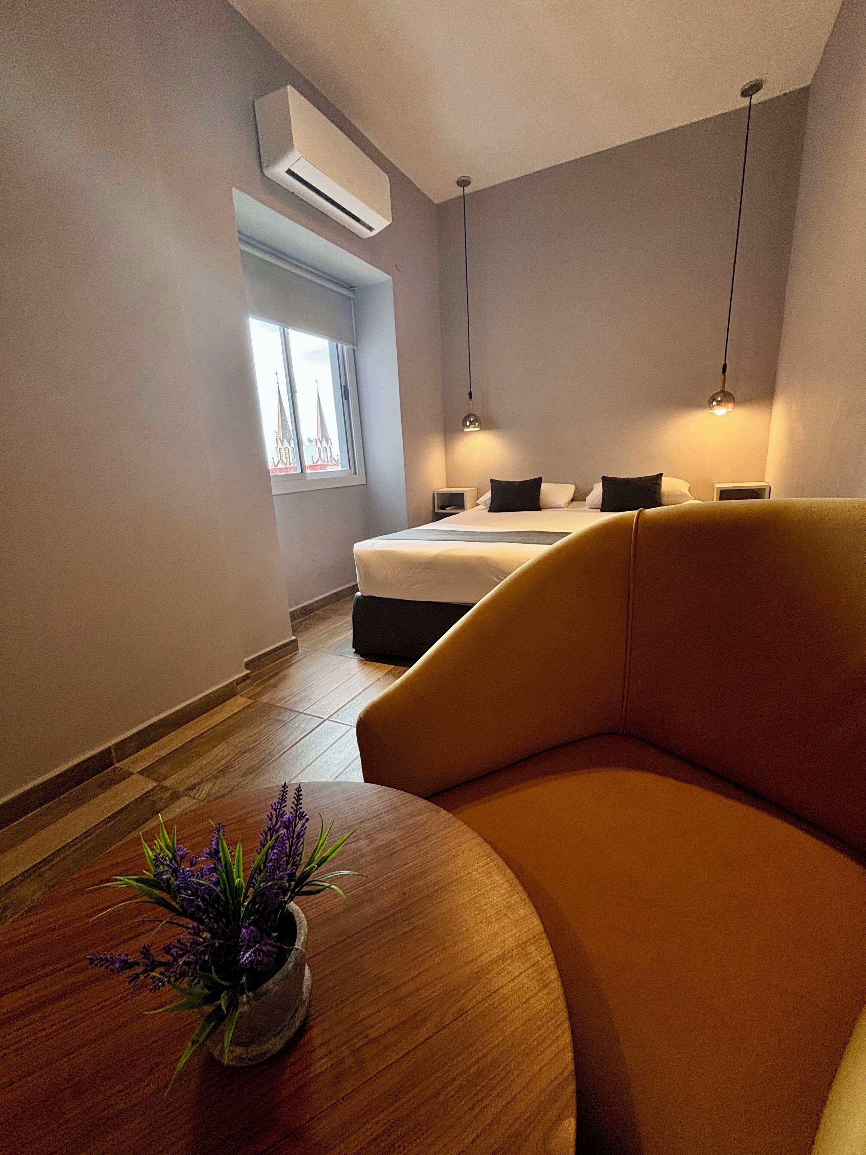 Une chambre d’hôtel moderne, équipée d’un lit, d’un canapé, d’une petite table avec une plante et d’une fenêtre donnant sur un immeuble.