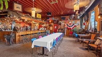Interior de un restaurante con una larga mesa común, zona de bar y diversas decoraciones, incluyendo banderas y un reloj.