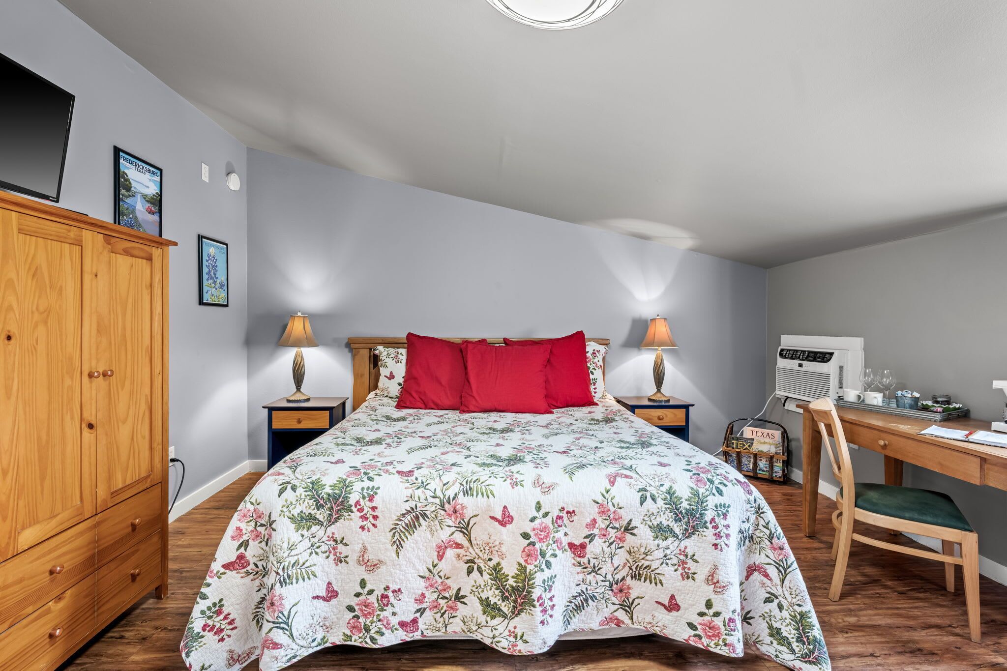 A bedroom with a floral bedspread, a wooden wardrobe, a desk with a chair, and a wall-mounted air conditioner.