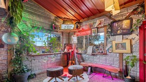 Une pièce chaleureuse au décor rustique, avec un plafond en bois, un mur en briques et une fenêtre offrant une belle vue. On y trouve deux tabourets, un banc et une guitare posés sur une table en bois.