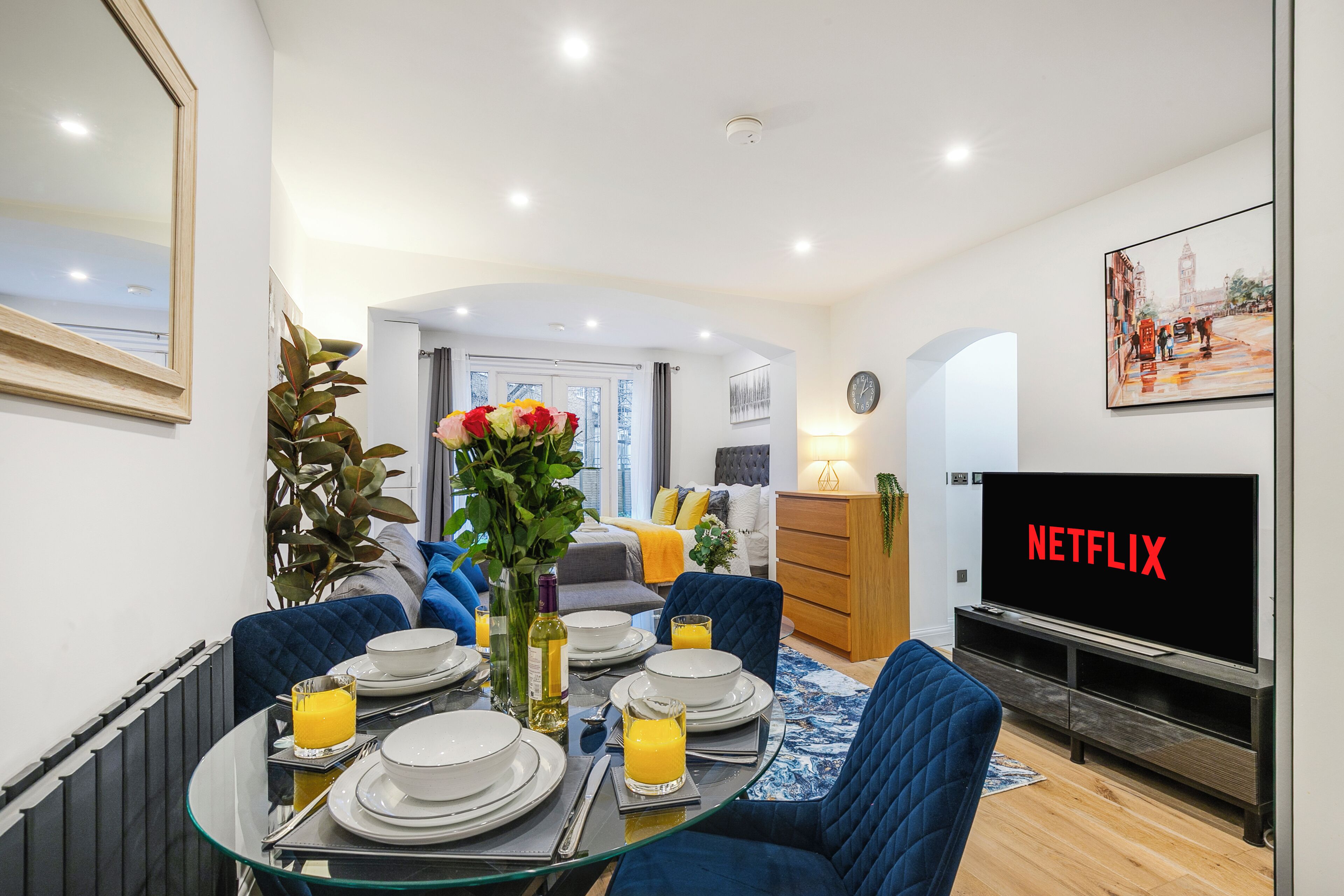 Een moderne woonkamer met een eetgedeelte, een tv waarop 'NETFLIX' wordt getoond, en een groot raam met uitzicht op de stad.