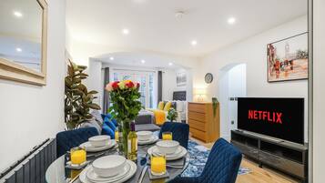 Een moderne woonkamer met een eetgedeelte, een tv waarop 'NETFLIX' wordt getoond, en een groot raam met uitzicht op de stad.