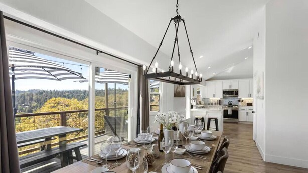 Comedor con mesa de madera dispuesta para seis, cocina al fondo y vista a árboles a través de amplias ventanas.