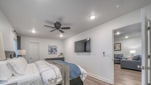 Un dormitorio con cama, ventilador de techo, televisor y una puerta que da a una sala de estar.
