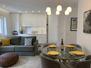 A modern living room with a glass dining table set for four, a grey sofa with cushions, a brown ottoman, and a kitchen in the background. - FLH Cascais Downtown Premium Apartment I (Cascais)