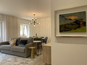 Un salon moderne avec un canapé gris, une petite table d’appoint ronde dorée et un coin repas avec une table dressée pour deux personnes.