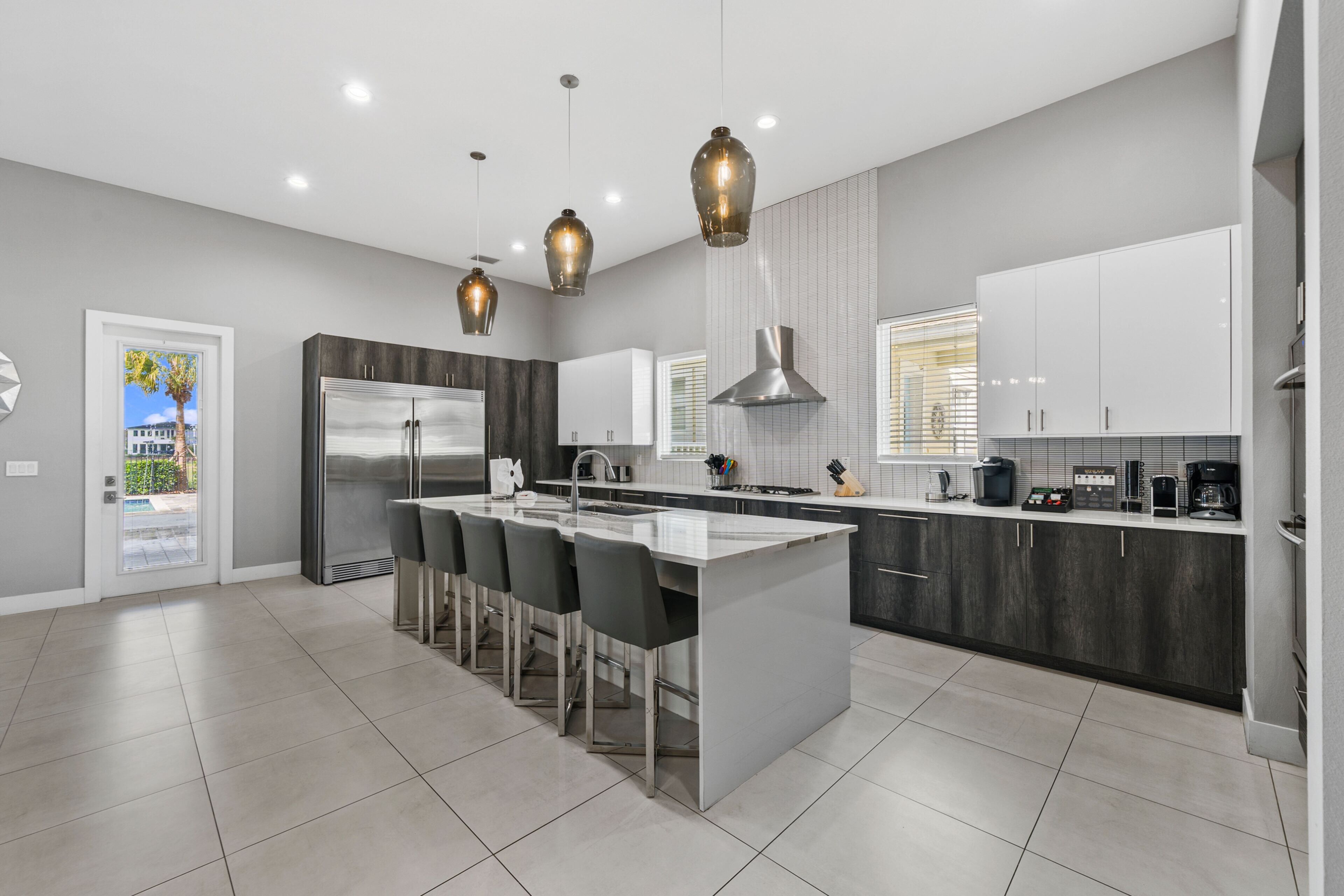 A modern kitchen with a central island, bar stools, stainless steel appliances, pendant lighting, and a view of the outside.