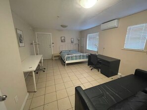 A small, well-lit room with a bed, a desk, a chair, and a window with blinds.