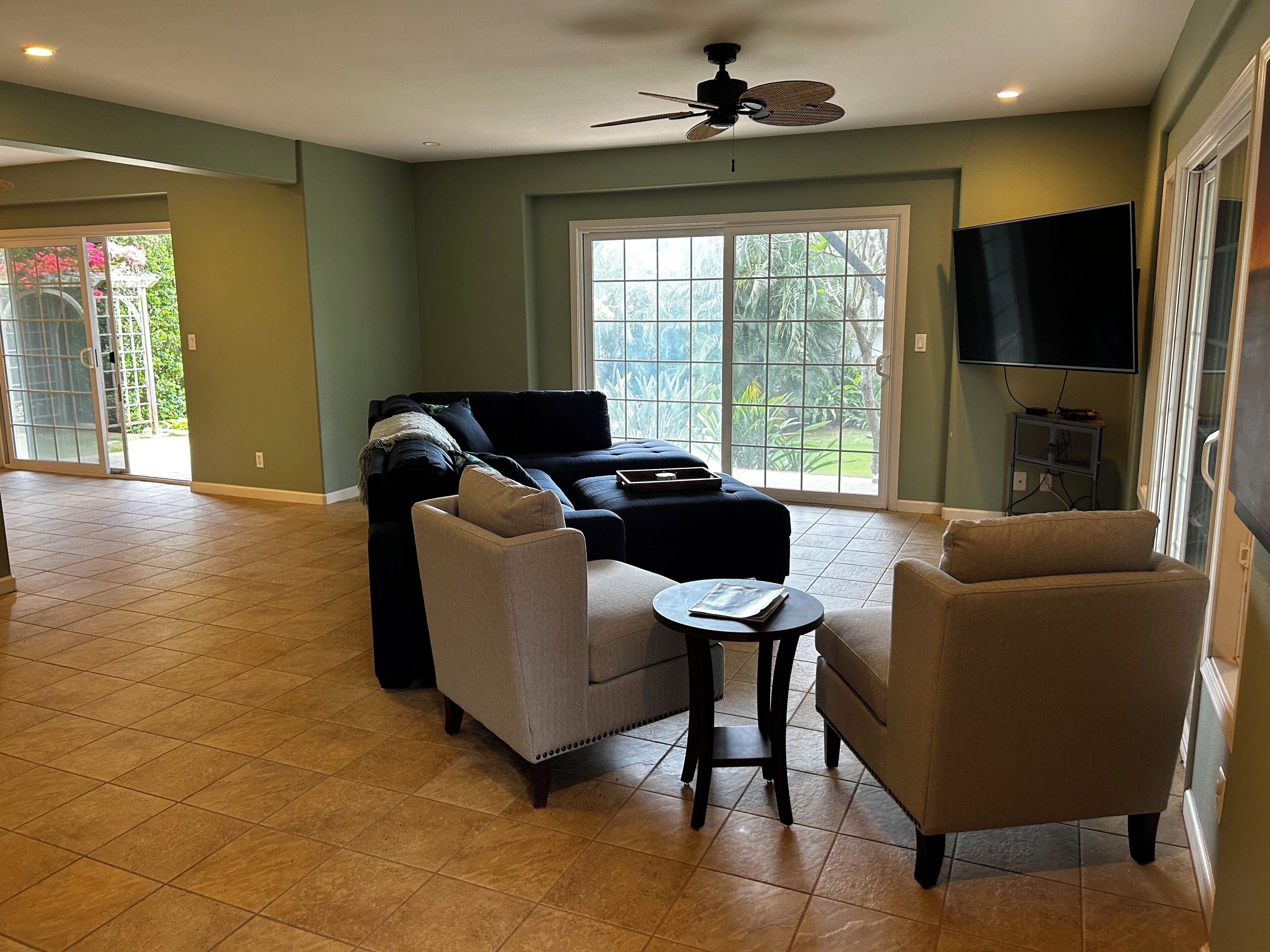 A spacious living room with a large window, a ceiling fan, and a flat-screen TV.