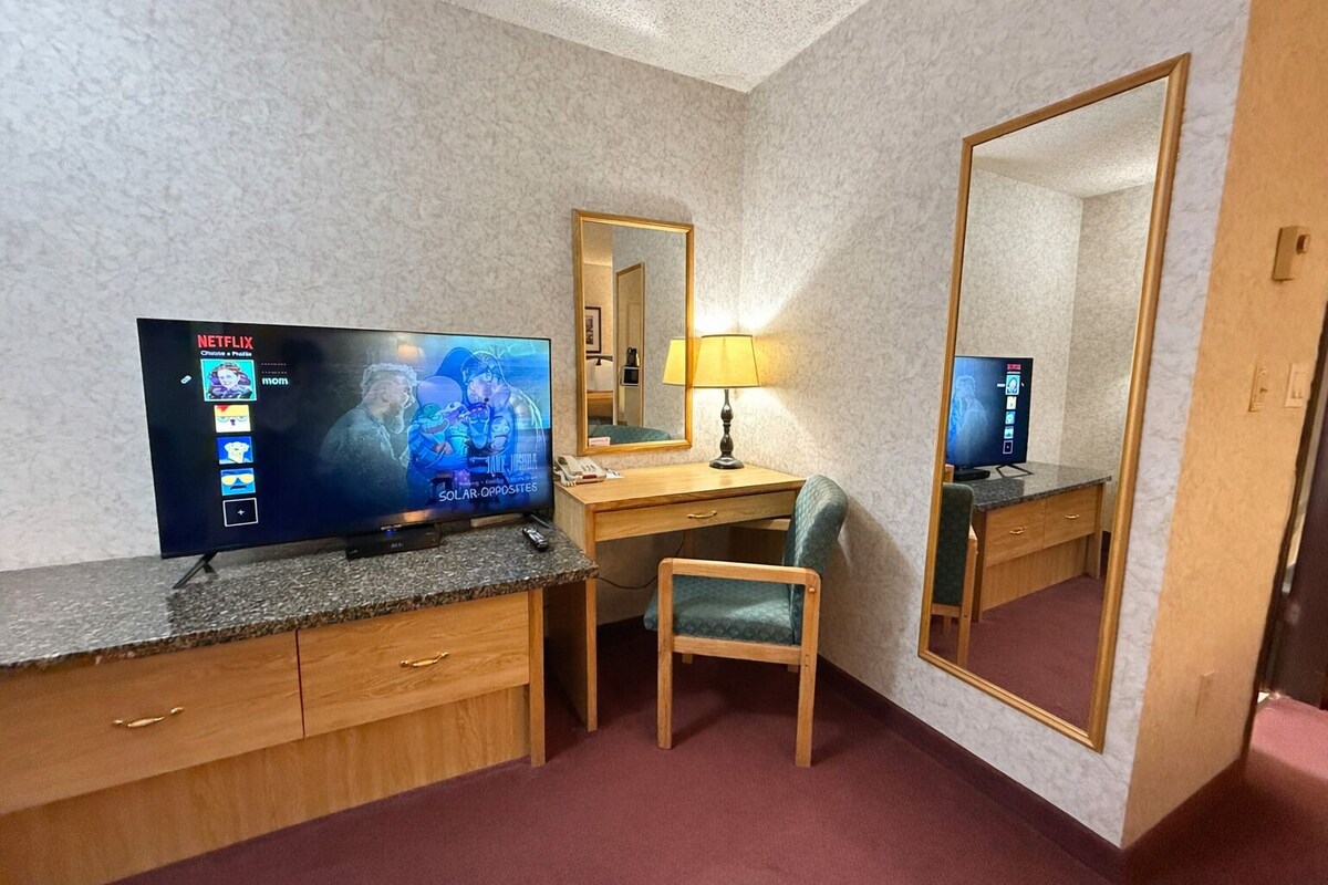 A hotel room with a TV displaying a Netflix interface, a desk with a lamp, a chair, and a large mirror.