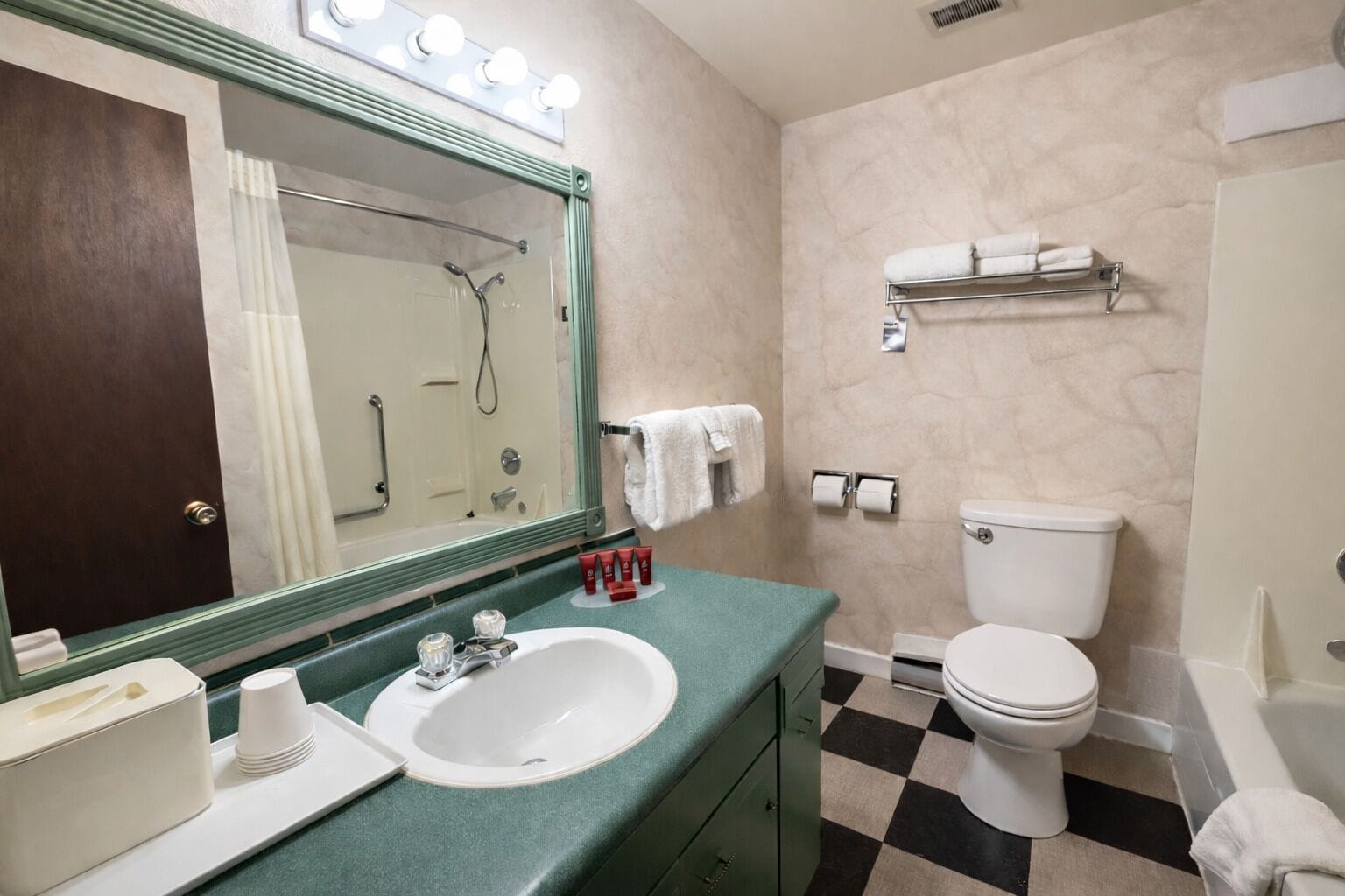 Un baño con lavamanos, espejo y retrete. Hay toallas en la pared y se ve una zona de ducha en el espejo.
