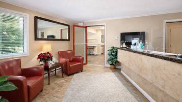 A hotel lobby with a reception desk, two red leather chairs, a small table with a lamp and a potted plant, and a large mirror on the wall.