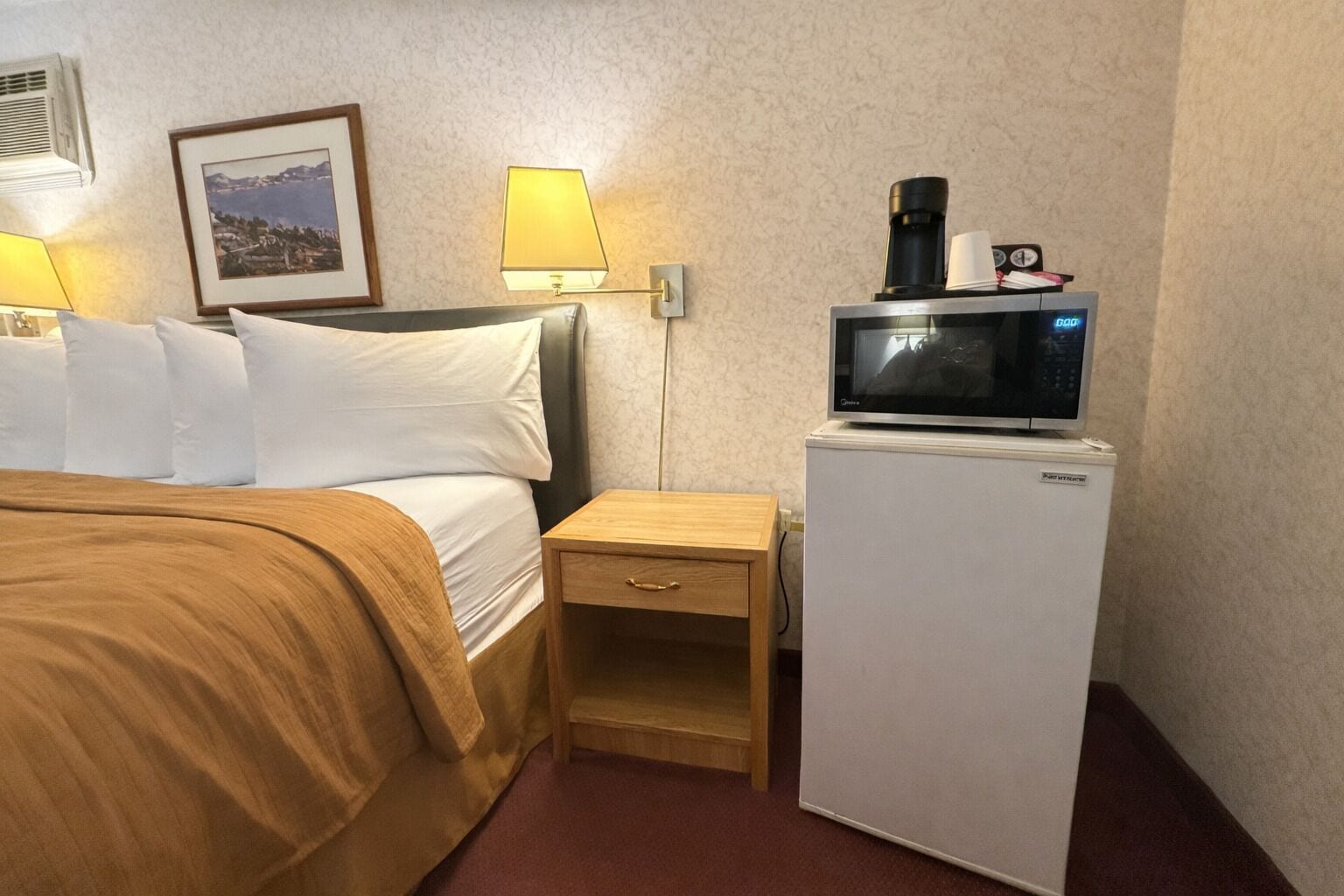 A hotel room with a bed, a nightstand, a small refrigerator, a microwave, a coffee maker, and a framed picture on the wall.