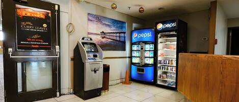 Un espace d’accueil comprenant un distributeur automatique de billets, un réfrigérateur rempli de produits Pepsi et un panneau indiquant le restaurant The Curry Leaves.